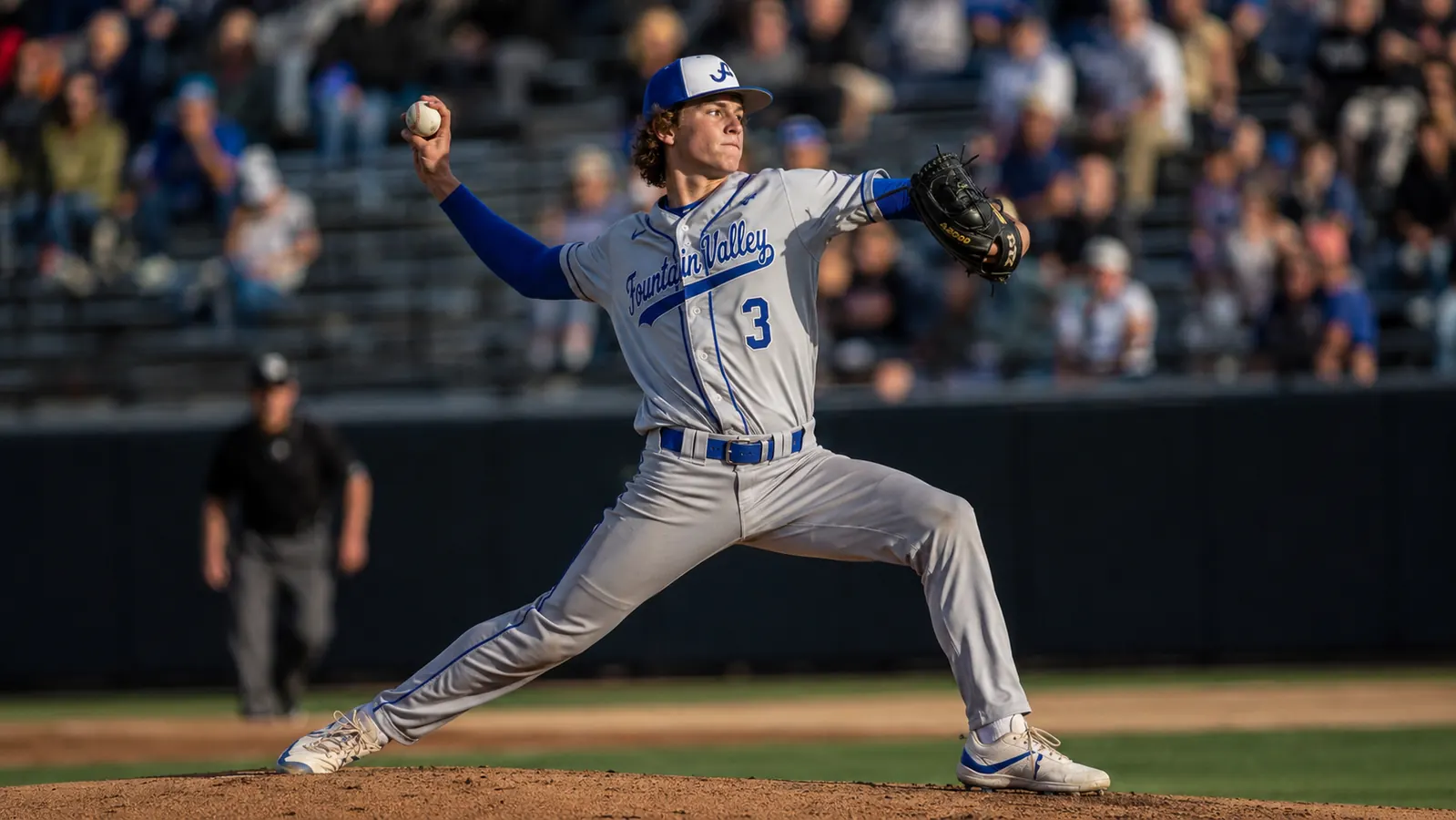 Teen pitcher throwing a pitch in a game, full wind-up visible, compression sleeve on throwing arm