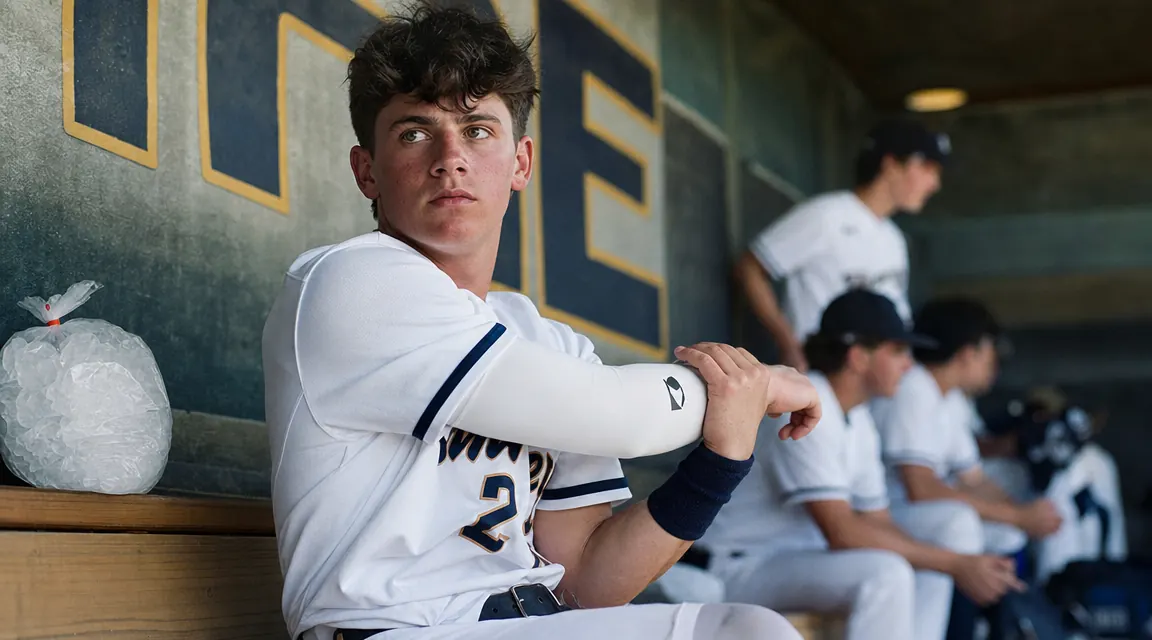 Teen pitcher in dugout stretching his pitching arm gently while an ice pack rests nearby on the bench