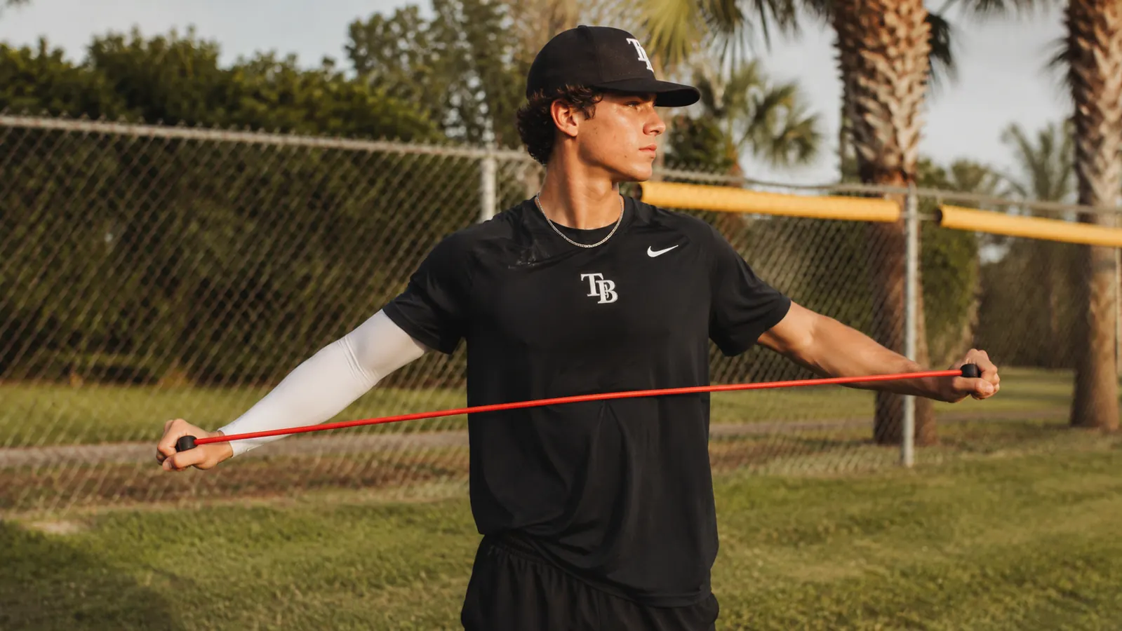 Teen pitcher doing light band exercises with Jaeger J-Bands outdoors before a game, compression sleeve visible on his throwing arm