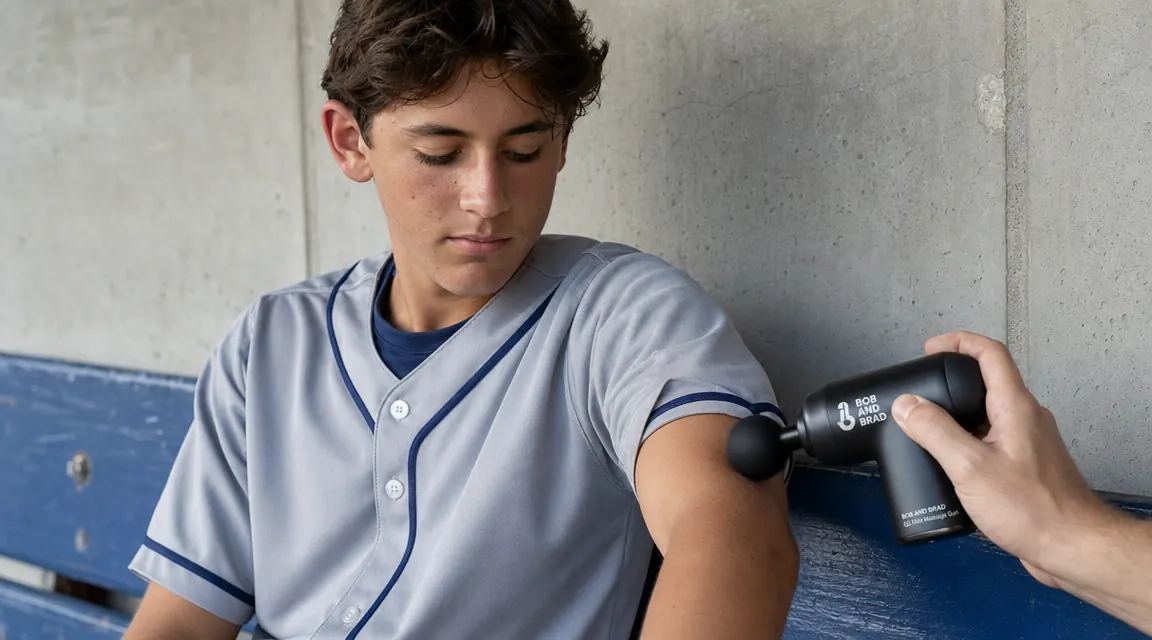 Hand holding a small black percussion massage gun against the forearm of a teen athlete sitting in dugout